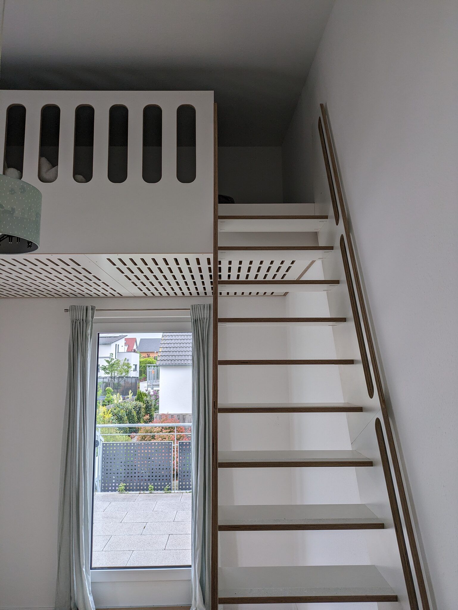 Weißes Hochbett unter der Decke eines Kinderzimmers: eine Brüstung mit länglichen Aussparungen, darunter eine gelochte Unterseite, rechts eine schräge Leiter mit Holzstufen.
