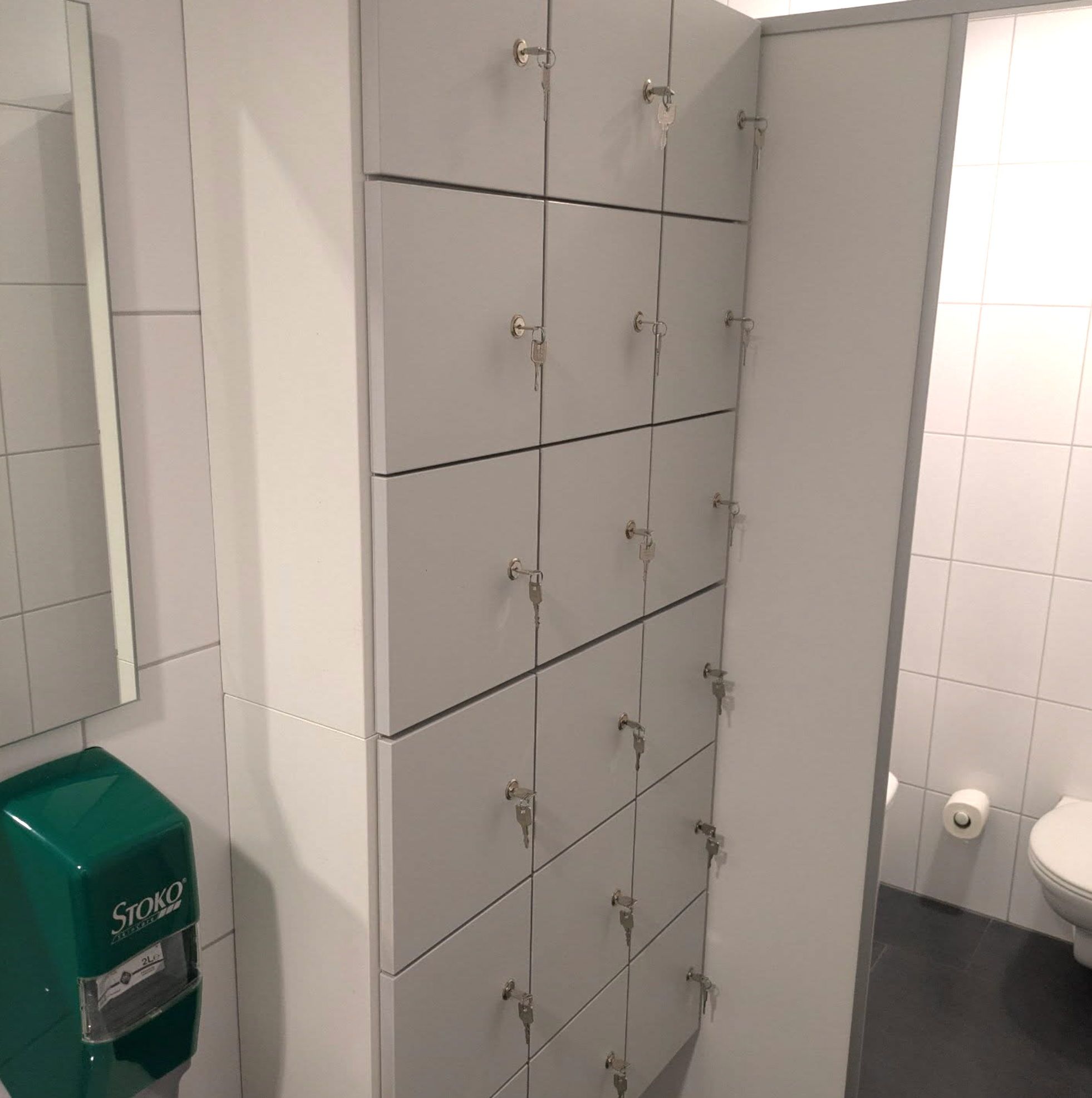 Weiße Schließfachschränke mit einzeln abschließbaren Türen, eingebaut in einen weiß gefliesten Sanitärraum.