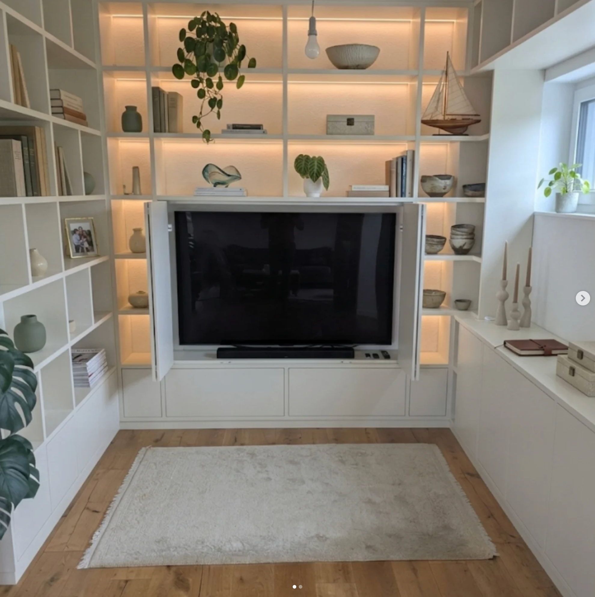 Wohnzimmer mit weißem Einbauregal über die ganze Wand: beleuchtete Fächer mit Büchern, Schalen und Pflanzen, in der Mitte ein Fernseher hinter geöffneten Falttüren, davor ein heller Teppich auf Eichenparkett.